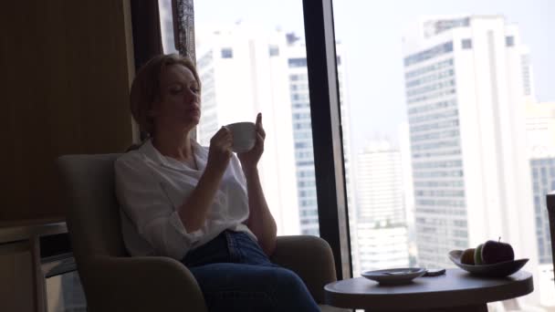 Hermosa mujer en una camisa blanca bebiendo café junto a la ventana con vistas a la ciudad. y utiliza su teléfono inteligente 4k — Vídeo de stock