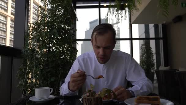 Um jovem empresário está tomando café da manhã no terraço do restaurante com vista para os arranha-céus. 4k . — Vídeo de Stock