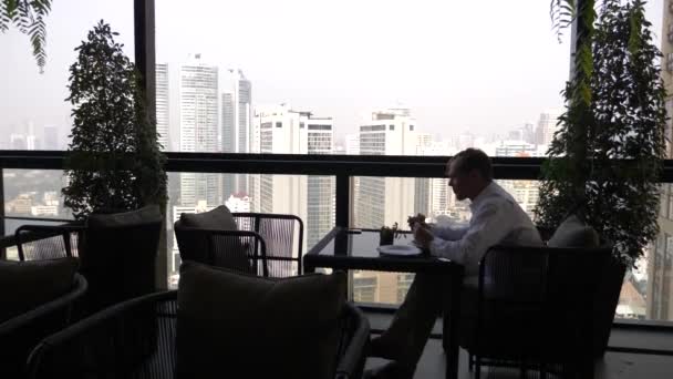 Młody biznesmen jest posiadanie śniadanie na tarasie restauracji z widokiem na wieżowce. 4k. — Wideo stockowe