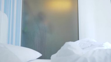 4k, ağır çekim. Tanınmayan kız duş alır. biri yatak odası ve banyo arasında şeffaf duvarı baktığı. Bulanık cam arkasındaki kadın. Kadın banyoda.