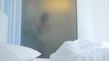 4k, ağır çekim. Tanınmayan kız duş alır. biri yatak odası ve banyo arasında şeffaf duvarı baktığı. Bulanık cam arkasındaki kadın. Kadın banyoda.