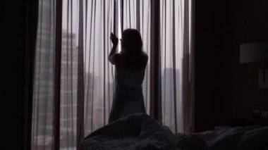 Bir kadın gökdelenler bakan bir pencere geliyor uykudan uyanır, perdeleri açar. ağır çekim. 4k