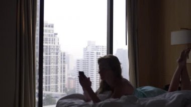 Kadın uykudan uyanır ve gökdelenler bir görünümü ile cama yatakta yatarken ona telefon kullanır. Ağır çekim. 4k