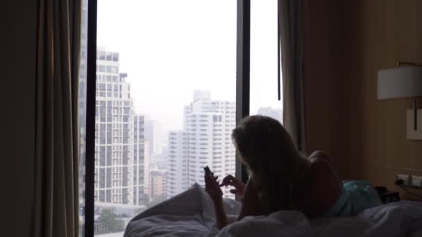 La donna si sveglia dal sonno e usa il suo telefono, sdraiata sul letto, contro la finestra con vista sui grattacieli. Al rallentatore. 4k — Video Stock