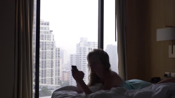 Wacht die Frau aus dem Schlaf auf und benutzt ihr Handy, auf dem Bett liegend, gegen das Fenster mit Blick auf die Wolkenkratzer. Zeitlupe. 4k — Stockvideo