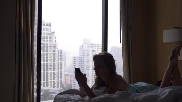 La mujer se despierta del sueño y usa su teléfono, acostada en la cama, contra la ventana con vista a los rascacielos. En cámara lenta. 4k — Vídeo de stock