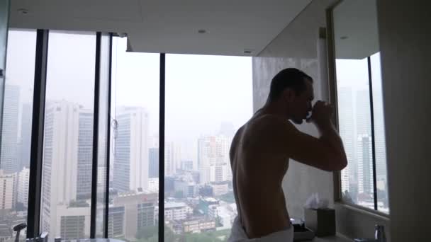 Young man, brushing teeth with a toothbrush in the bathroom with a panoramic window. view from the window to the skyscrapers. 4k, slow motion — Stock Video