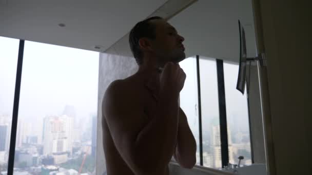 Um homem bonito aplicando uma navalha no banheiro com vista da janela para os arranha-céus. 4k, câmera lenta — Vídeo de Stock