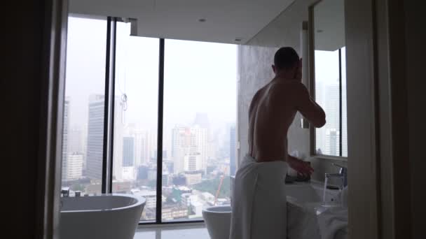 Un bell'uomo che applica un rasoio in bagno con vista dalla finestra ai grattacieli. 4k, rallentatore — Video Stock
