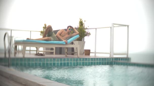 Un hombre relajado, tomando el sol en las tumbonas cerca de la piscina en el techo con vistas al mar. 4k, desenfoque de fondo — Vídeos de Stock