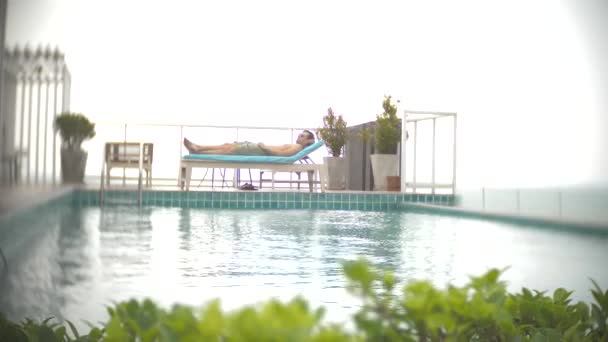 Ένας άνδρας χαλάρωση, ηλιοθεραπεία στις ξαπλώστρες δίπλα στην πισίνα στην ταράτσα με θέα στη θάλασσα. 4k, θάμπωμα του φόντου — Αρχείο Βίντεο