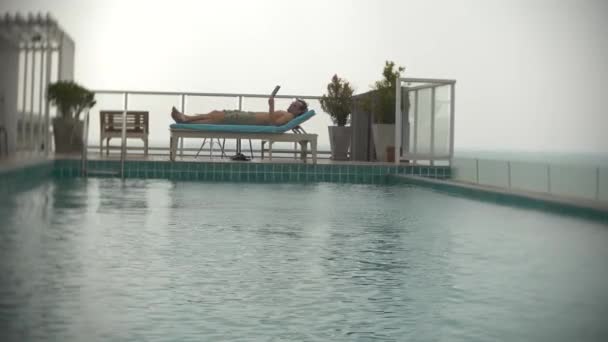 Un joven con una tableta en la piscina en el techo con vistas al mar. 4k. , difuminar el fondo — Vídeos de Stock
