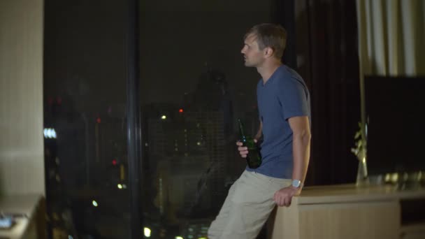 Мужчина пьет пиво дома рядом с панорамным окном с видом на небоскребы. ночь, фоновое размытие. 4k, slow motion — стоковое видео