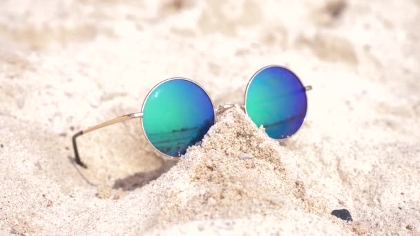 Солнцезащитные очки лежат на песке на пляже. Солнечные очки отражают море, солнце, небо, пляж. 4k, slow motion — стоковое видео