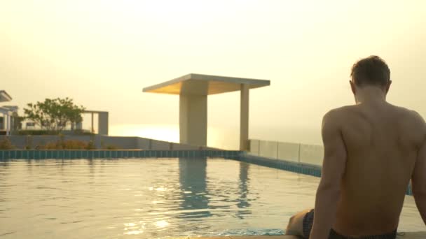 O homem senta-se na borda da piscina e molha os pés na água. uma piscina luxuosa no telhado da casa com vista para o mar. Ao pôr-do-sol. 4k, câmera lenta, brilho solar na água . — Vídeo de Stock