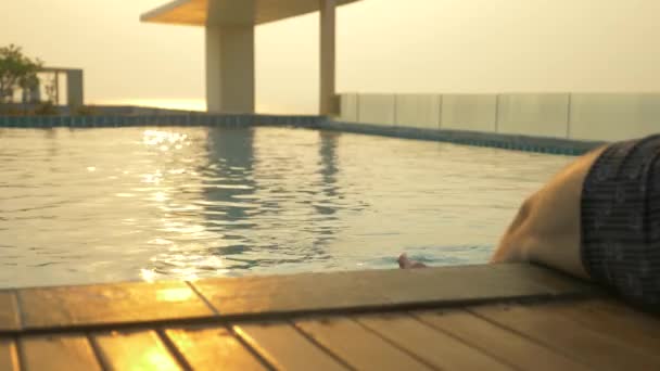 男はプールの端に座っているし、水に彼の足を濡らします。海の見える家の屋根の上の豪華なプール。夕日。4 k、スローモーション、水面に太陽のまぶしさ. — ストック動画