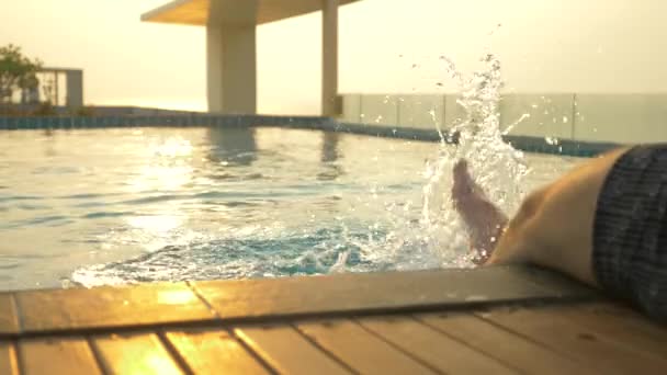 L'uomo siede sul bordo della piscina e bagna i piedi nell'acqua. una lussuosa piscina sul tetto della casa con vista sul mare. Al tramonto. 4k, rallentatore, bagliore solare sull'acqua . — Video Stock