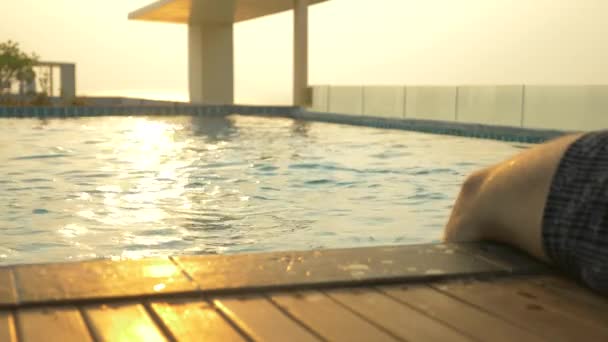 Sitzt der Mann am Beckenrand und benetzt seine Füße im Wasser. ein luxuriöser Pool auf dem Dach des Hauses mit Meerblick. am Sonnenuntergang. 4k, Zeitlupe, Sonnenblendung auf dem Wasser. — Stockvideo