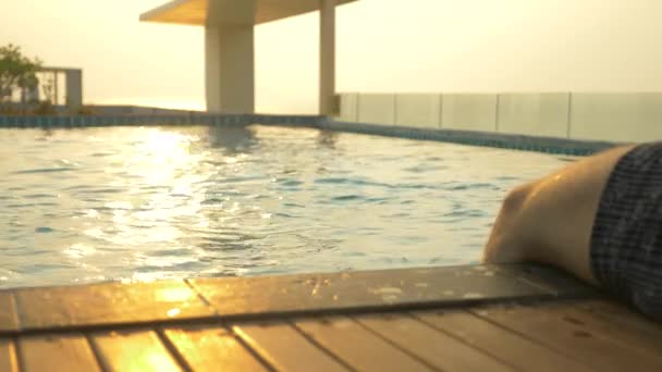 L'uomo siede sul bordo della piscina e bagna i piedi nell'acqua. una lussuosa piscina sul tetto della casa con vista sul mare. Al tramonto. 4k, rallentatore, bagliore solare sull'acqua . — Video Stock