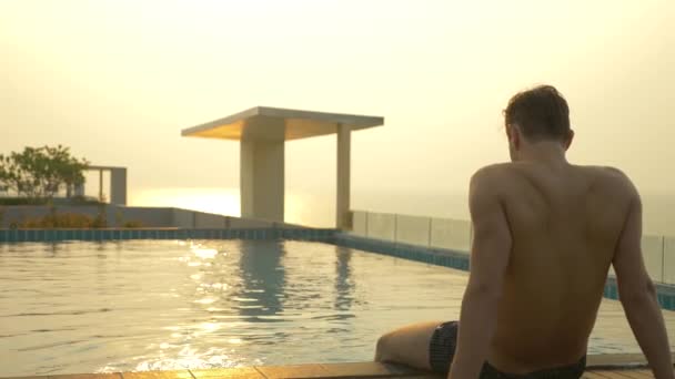El hombre se sienta en el borde de la piscina y moja sus pies en el agua. una lujosa piscina en el techo de la casa con vistas al mar. En la puesta del sol. 4k, cámara lenta, deslumbramiento solar en el agua . — Vídeo de stock