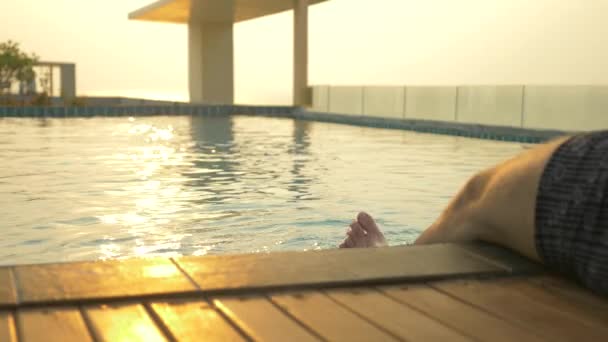 O homem senta-se na borda da piscina e molha os pés na água. uma piscina luxuosa no telhado da casa com vista para o mar. Ao pôr-do-sol. 4k, câmera lenta, brilho solar na água . — Vídeo de Stock