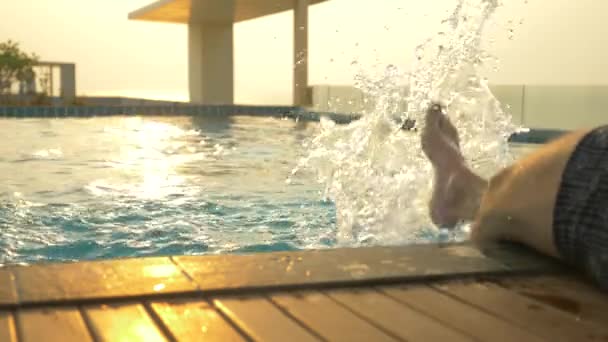 男はプールの端に座っているし、水に彼の足を濡らします。海の見える家の屋根の上の豪華なプール。夕日。4 k、スローモーション、水面に太陽のまぶしさ. — ストック動画