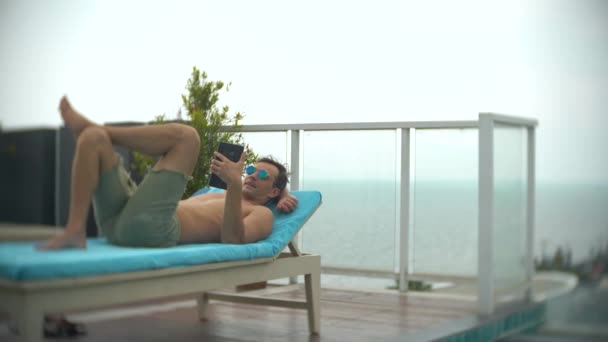 Ein junger Mann mit einem Tablet am Pool auf dem Dach mit Blick aufs Meer. 4k. , den Hintergrund verwischen — Stockvideo