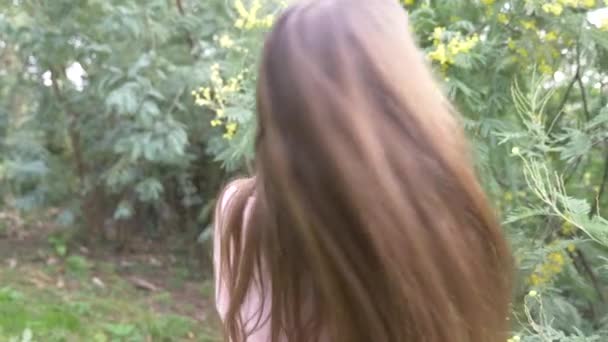 Chica adolescente endereza su pelo largo. Ella está junto al árbol de mimosa floreciente. 4k, cámara lenta — Vídeo de stock