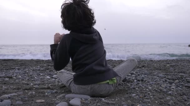 Un chico solitario se sienta en la orilla y arroja piedras al mar. 4k, cámara lenta — Vídeo de stock