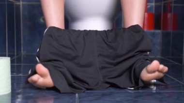 onun midesinde bir acıyla tuvalette oturan bir adamın ayakları. 4k, yakın çekim, tuvalette şortlu genç adam.