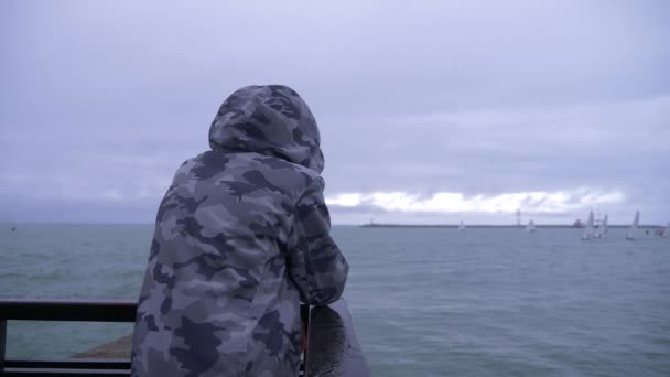 Alguien con una chaqueta encapuchada está de pie en el muelle y viendo los veleros navegando hacia el mar. 4k, cámara lenta . — Vídeo de stock