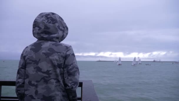 Alguien con una chaqueta encapuchada está de pie en el muelle y viendo los veleros navegando hacia el mar. 4k, cámara lenta . — Vídeo de stock