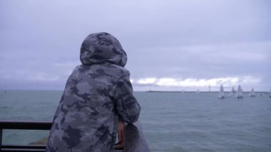 birisi kapşonlu ceket iskelede duran ve denize yelken yelkenli izliyor. 4k, ağır çekim.