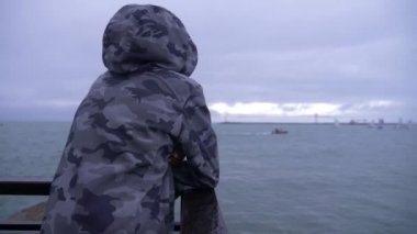 birisi kapşonlu ceket iskelede duran ve denize yelken yelkenli izliyor. 4k, ağır çekim.