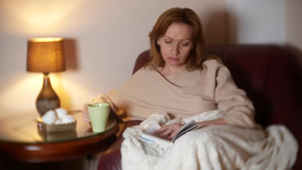 Por la noche, una chica está leyendo un libro debajo de una manta en una silla. bebiendo té caliente y comiendo malvaviscos. El concepto de relajación, la lectura de un libro. 4k, borra el borde del marco — Vídeo de stock