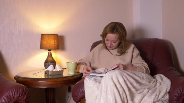 Por la noche, una chica está leyendo un libro debajo de una manta en una silla. bebiendo té caliente y comiendo malvaviscos. sonríe y se ríe de lo que lee en el libro 4k, cámara lenta — Vídeos de Stock