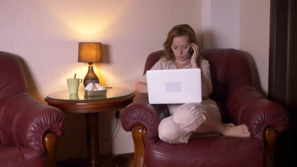 Mujer que trabaja de noche en el ordenador portátil y comer bocadillos, sentado en sillón en casa. 4k, cámara lenta . — Vídeos de Stock