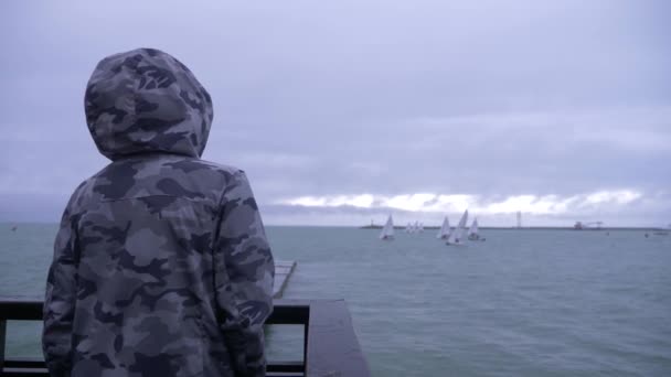 有人穿着带头巾的夹克站在码头上, 看着帆船驶向大海。4k. 慢动作. — 图库视频影像