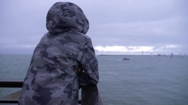 Кто-то в капюшоне стоит на пирсе и наблюдает за парусниками, плывущими к морю. 4k, slow motion . — стоковое видео