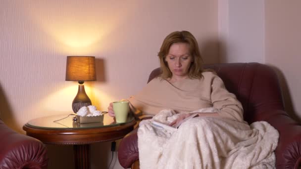 Di notte, una ragazza sta leggendo un libro sotto una coperta su una sedia. sorseggiando tè caldo e mangiando marshmallow. Il concetto di relax, leggere un libro. 4k, rallentatore — Video Stock