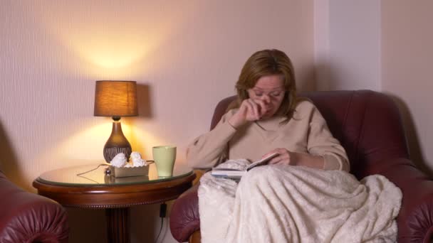 Di notte, una ragazza sta leggendo un libro sotto una coperta su una sedia. sorseggiando tè caldo e mangiando marshmallow. sorride e ride di quello che ha letto nel libro 4k, slow motion — Video Stock