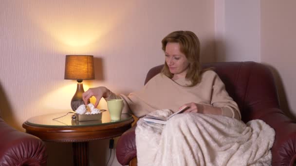 v noci dívka čte knihu pod deku na židli. usrkával horký čaj a jíst marshmallows. Pojem relaxace, čtení knihy. 4k, pomalý pohyb