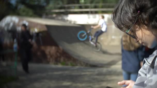 Een jongen die alleen met behulp van een mobiele telefoon in de buitenlucht. terwijl andere kinderen actieve rust hebben. Smartphone verslaving. 4k, slow-motion — Stockvideo