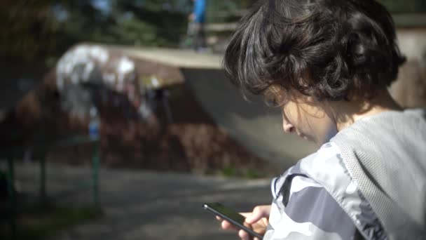 Een jongen die alleen met behulp van een mobiele telefoon in de buitenlucht. terwijl andere kinderen actieve rust hebben. Smartphone verslaving. 4k, slow-motion — Stockvideo