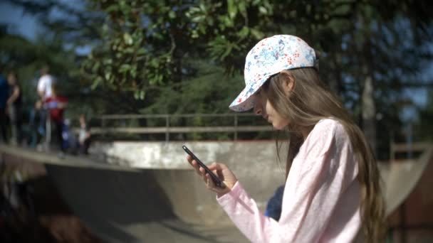 Una chica sola usando un teléfono móvil al aire libre. mientras otros niños descansan activamente. Adicción a teléfonos inteligentes. 4k, cámara lenta — Vídeo de stock