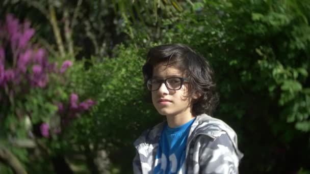 Porträtt av en pojke som bär glasögon, tittar på kameran i en park i sommar. 4 k. slowmotion — Stockvideo