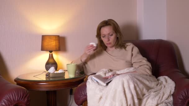 À noite, uma menina está lendo um livro debaixo de um cobertor em uma cadeira. Beber chá quente e comer marshmallows. O conceito de relaxamento, a leitura de um livro. 4k, câmera lenta — Vídeo de Stock