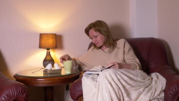 Por la noche, una chica está leyendo un libro debajo de una manta en una silla. bebiendo té caliente y comiendo malvaviscos. sonríe y se ríe de lo que lee en el libro 4k, cámara lenta — Vídeos de Stock