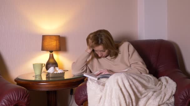 Por la noche la muchacha lee el libro bajo la manta en la silla. se queda dormida leyendo un libro aburrido no interesante de 4k, cámara lenta — Vídeos de Stock