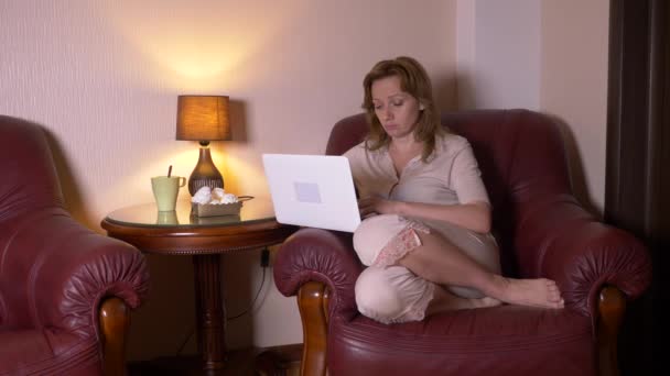 Mulher trabalhando à noite no laptop e comer lanches, sentado em poltrona em casa. 4k, câmera lenta . — Vídeo de Stock